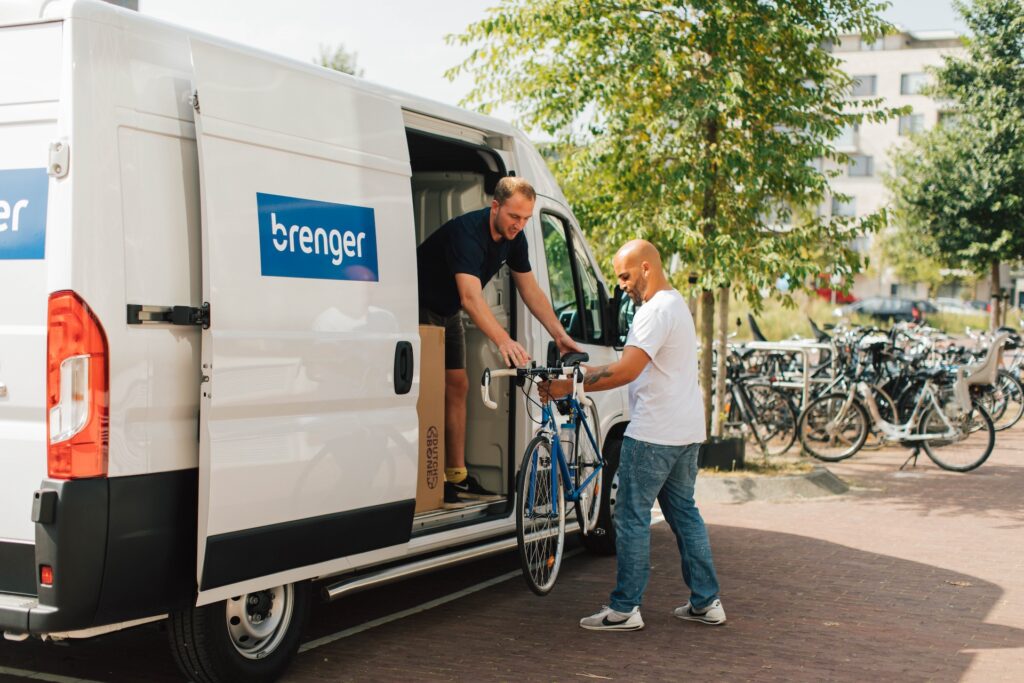 Twee mannen laden een blauwe racefiets uit een witte bestelbus met Brenger-logo. De ene man staat in de bus terwijl de andere man de fiets aanneemt. Op de achtergrond zijn fietsenstallingen en groen zichtbaar. Ideaal voor het duurzaam en efficiënt bezorgen van fietsen.