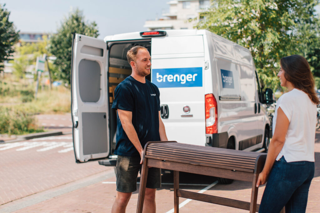 Een man en vrouw tillen samen een houten meubelstuk uit een Brenger-busje. De bestelbus met Brenger-logo staat open geparkeerd in een groene woonomgeving. Ideaal voor het huren van een busje voor duurzaam en efficiënt transport.