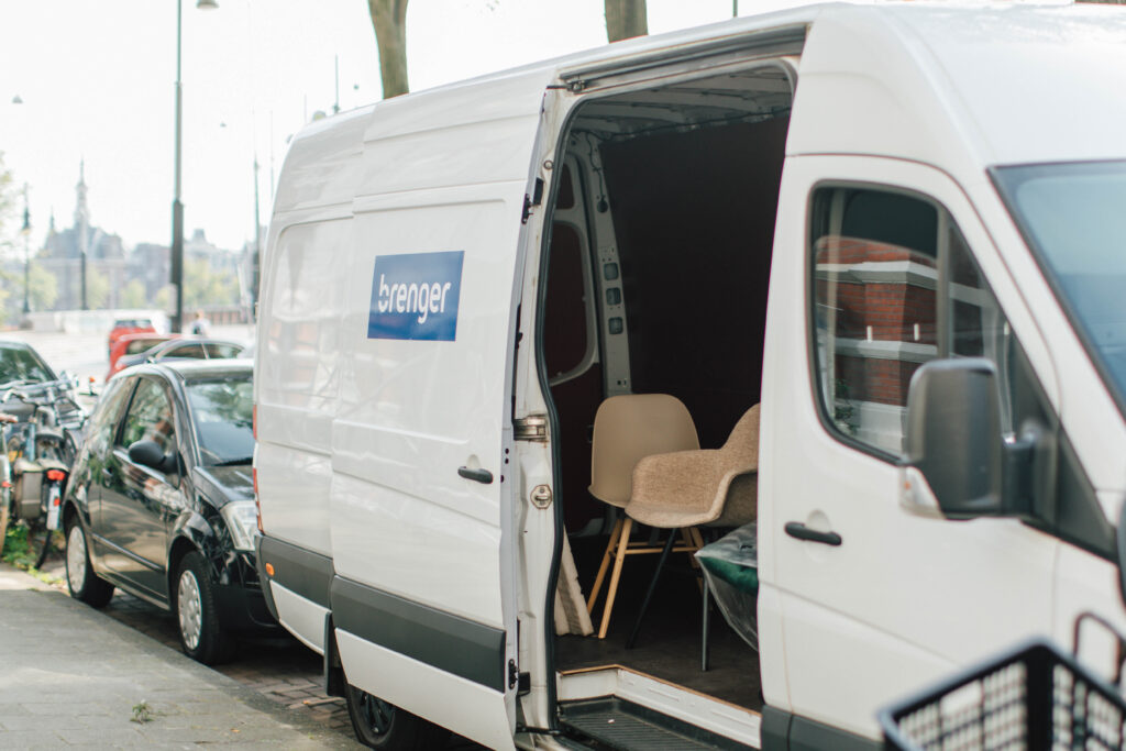 Een witte bestelbus met Brenger-logo staat geparkeerd aan de straatkant. De schuifdeur van de bus staat open, waardoor enkele stoelen zichtbaar zijn die binnenin staan, klaar om meubels duurzaam te verzenden. Op de achtergrond staan auto's, fietsen en gebouwen.