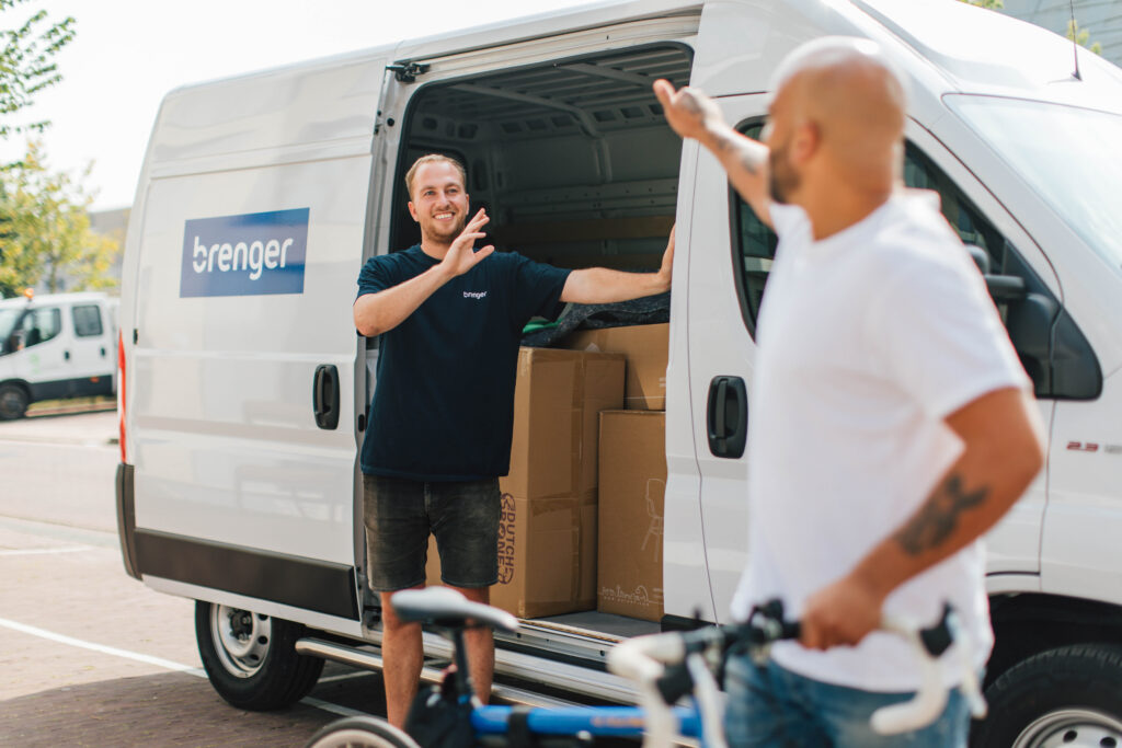 Een Brenger-koerier staat bij een witte bestelbus vol grote pakketten en zwaait naar een klant met een fiets. De bestelbus, voorzien van het Brenger-logo, benadrukt de efficiënte en duurzame manier van verzenden van grote pakketten. Op de achtergrond zijn meer voertuigen en een groene omgeving zichtbaar.
